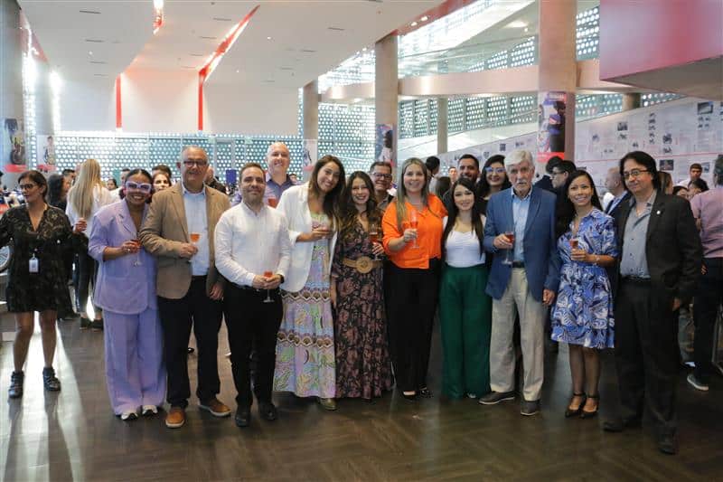 Grupo grande de personas, aproximadamente 16, posando para una foto de grupo en un espacio interior amplio y moderno. **Contenido principal:** El foco principal es el grupo de personas, de diversas edades y etnias, vestidas con ropa de cóctel informal. Parece ser un evento social o una reunión, dado el ambiente relajado y la proximidad del grupo. Hay una mezcla de hombres y mujeres, con una distribución bastante equitativa.