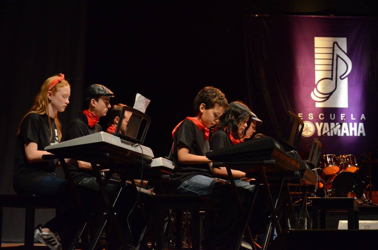 niños y adolescentes tocando teclados eléctricos en un escenario. Todos están vestidos con camisetas negras y llevan pañuelos rojos alrededor del cuello, lo que les da un toque uniforme. Están sentados frente a los teclados, muy concentrados en su ejecución musical. En primer plano, se destaca una niña con cabello largo y rojo, tocando un teclado, mientras que los otros niños y adolescentes están igualmente enfocados en sus instrumentos. Al fondo, se ve una gran pancarta con el logo de la 