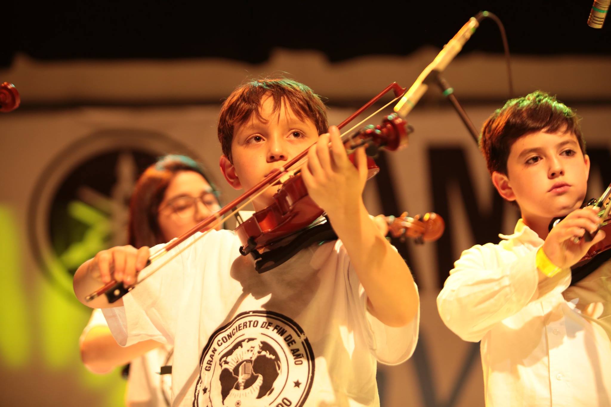 La imagen muestra a dos niños tocando el violín en un escenario, durante lo que parece ser una presentación en vivo. Ambos niños están concentrados en su música, con el violín en sus manos y el arco en movimiento. El niño de la izquierda, que tiene el cabello castaño claro, usa una camiseta blanca con un logo que dice 