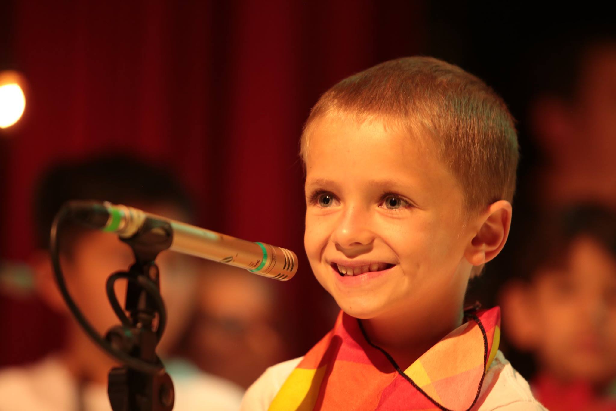 Niño sonriendo frente a microfono con un pañuelo en su cuello de colores en tonos rojos y naranjas