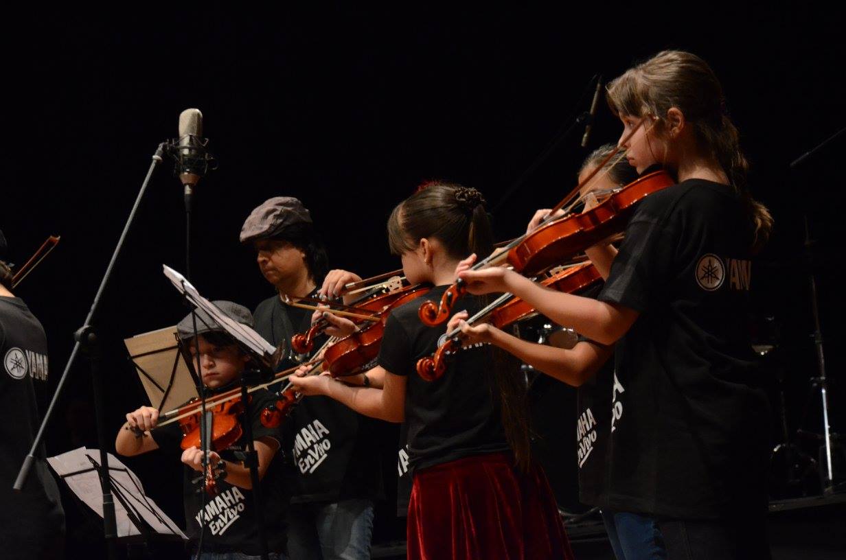 grupo de niños y adolescentes tocando el violín en un escenario, probablemente durante una presentación en vivo. Están vestidos con camisetas negras con el logotipo de Yamaha en la parte frontal. Los niños están concentrados, tocando sus instrumentos, algunos con partituras frente a ellos, mientras otros tienen el rostro serio y enfocado en la música. En primer plano, se puede ver a una niña con cabello recogido y una falda roja, tocando su violín con mucha dedicación. En el fondo, hay un micrófono de escenario y varios otros niños también tocando violín